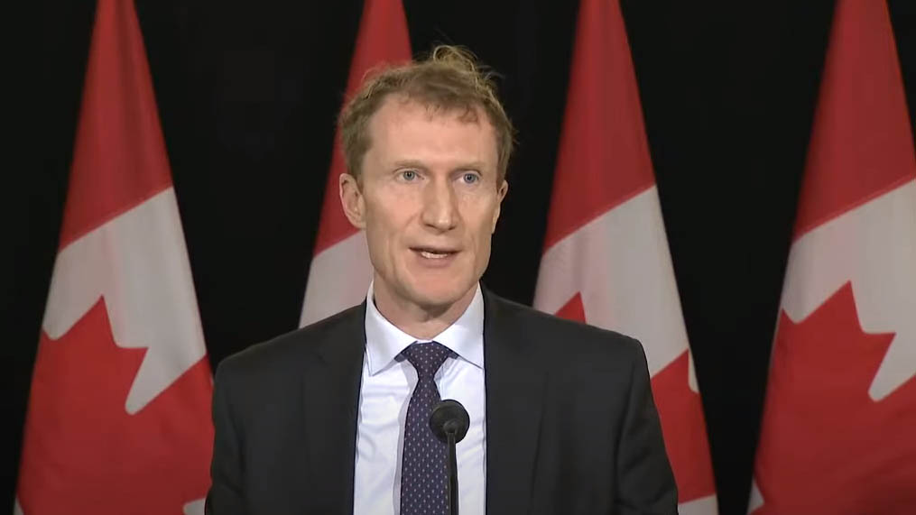 Marc Miller standing in front of a microphone. Canadian flags are shown in the background.
