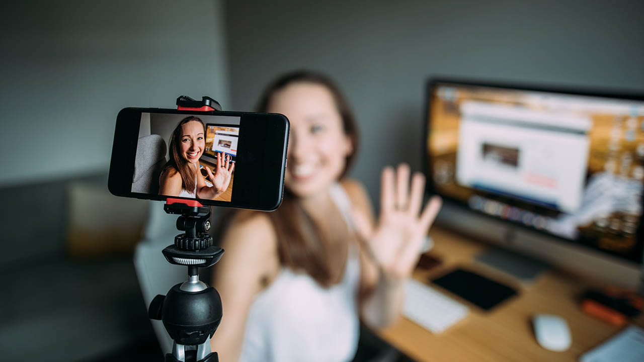 A person is seen recording themselves using a selfie stick and phone. The person is waving. A computer is shown behind the person.