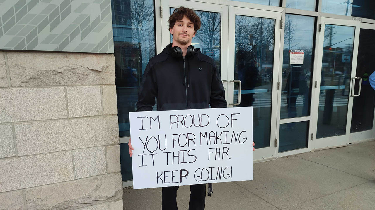 Dylan Dugal holding a sign outside a building. The sign states: I'm proud of you for making it this far. Keep going!