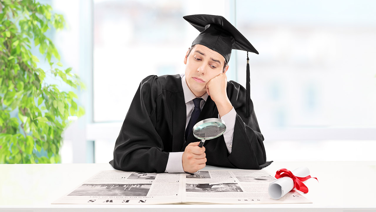 Stock photo of a young man in graduation a cap and gown, holding a magnifying glass as he looks for jobs.