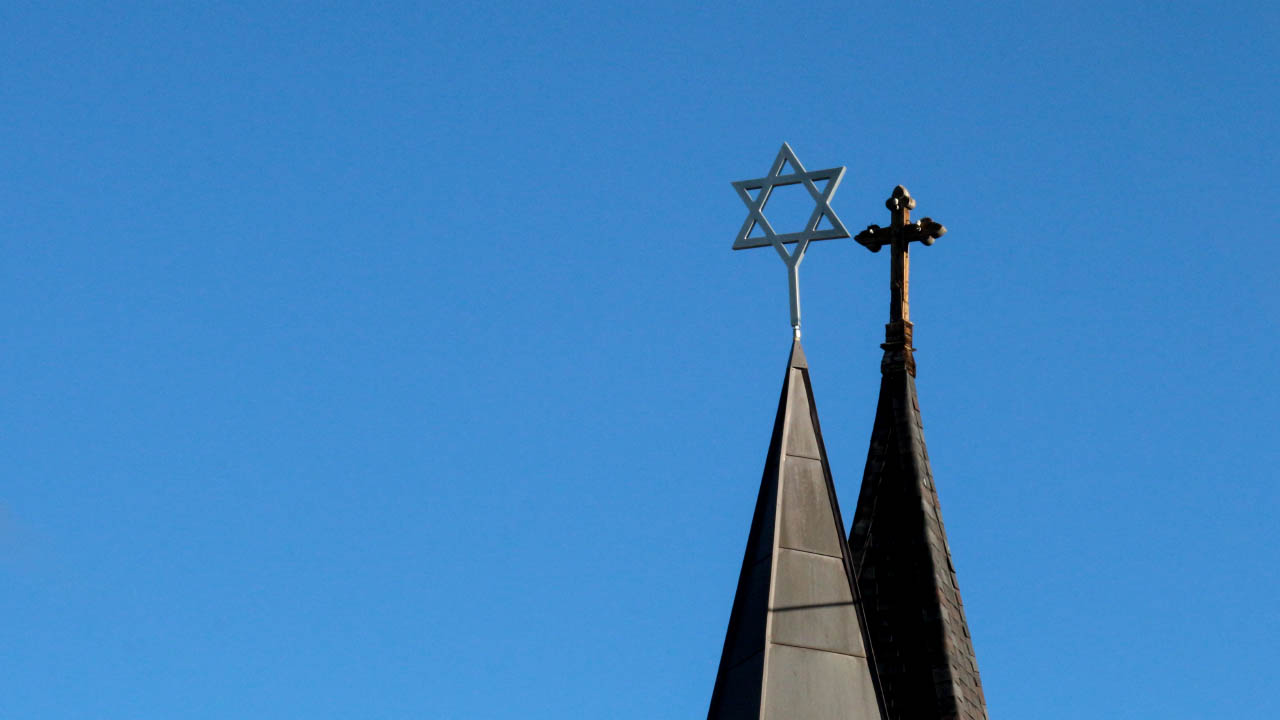 An image of two church steeples. The closer one has the star of David, the further one has a cross.