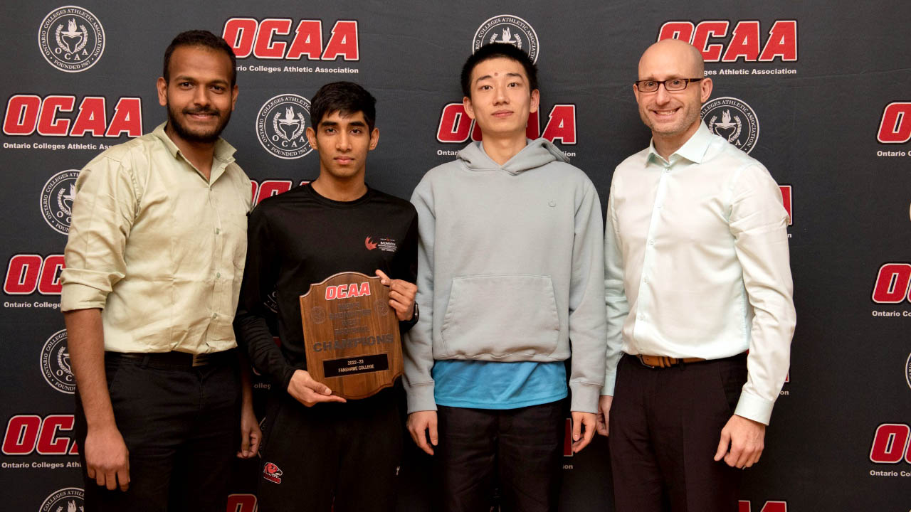 Four individuals pose in front of a backdrop with OCAA logos on it. One is holding a plaque that says OCAA champions.
