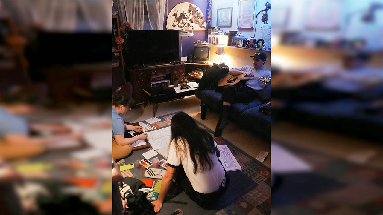 A photo of artists painting on the floor while another artist sits on a couch with a guitar. 