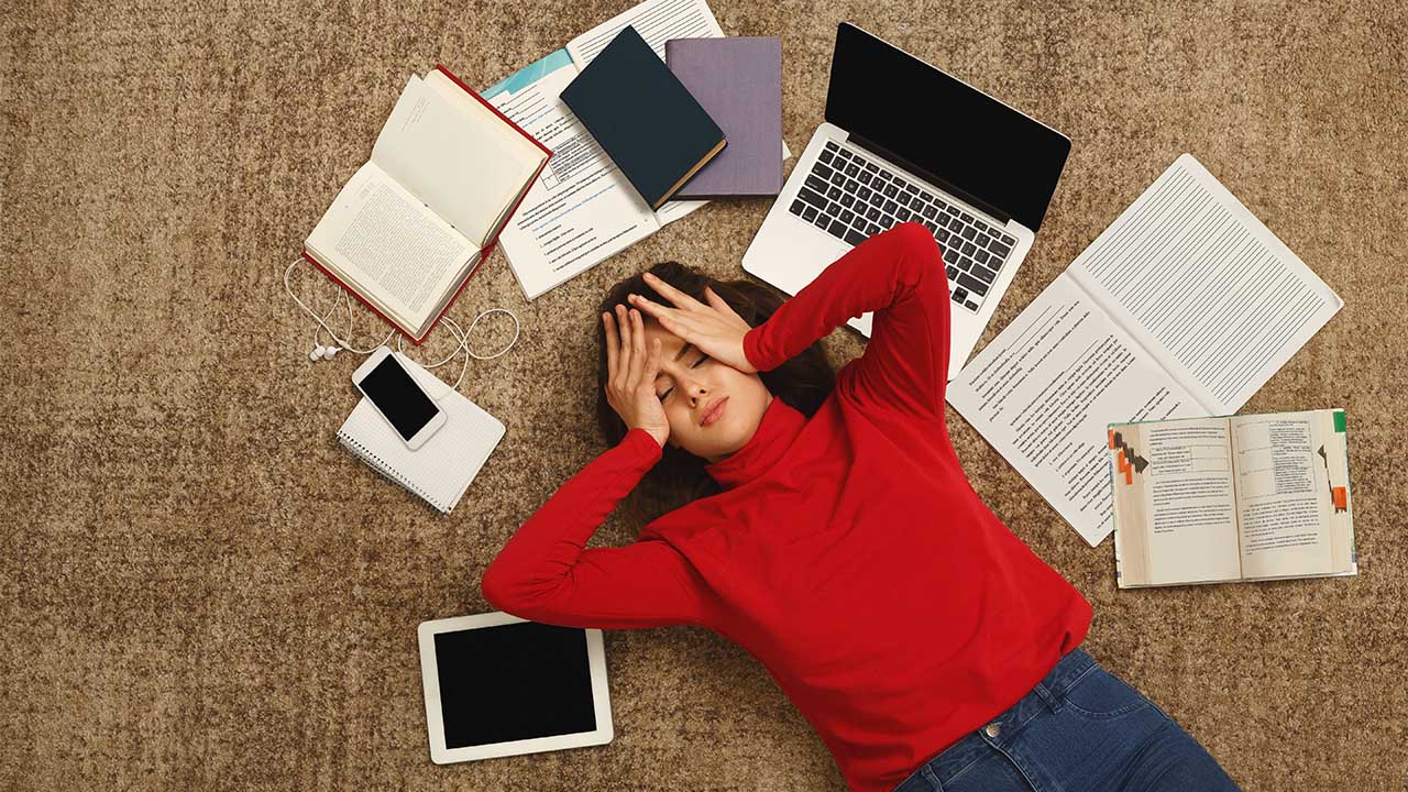 Stock image of a student lying stressed out on the floor surrounded by textbooks and a laptop.